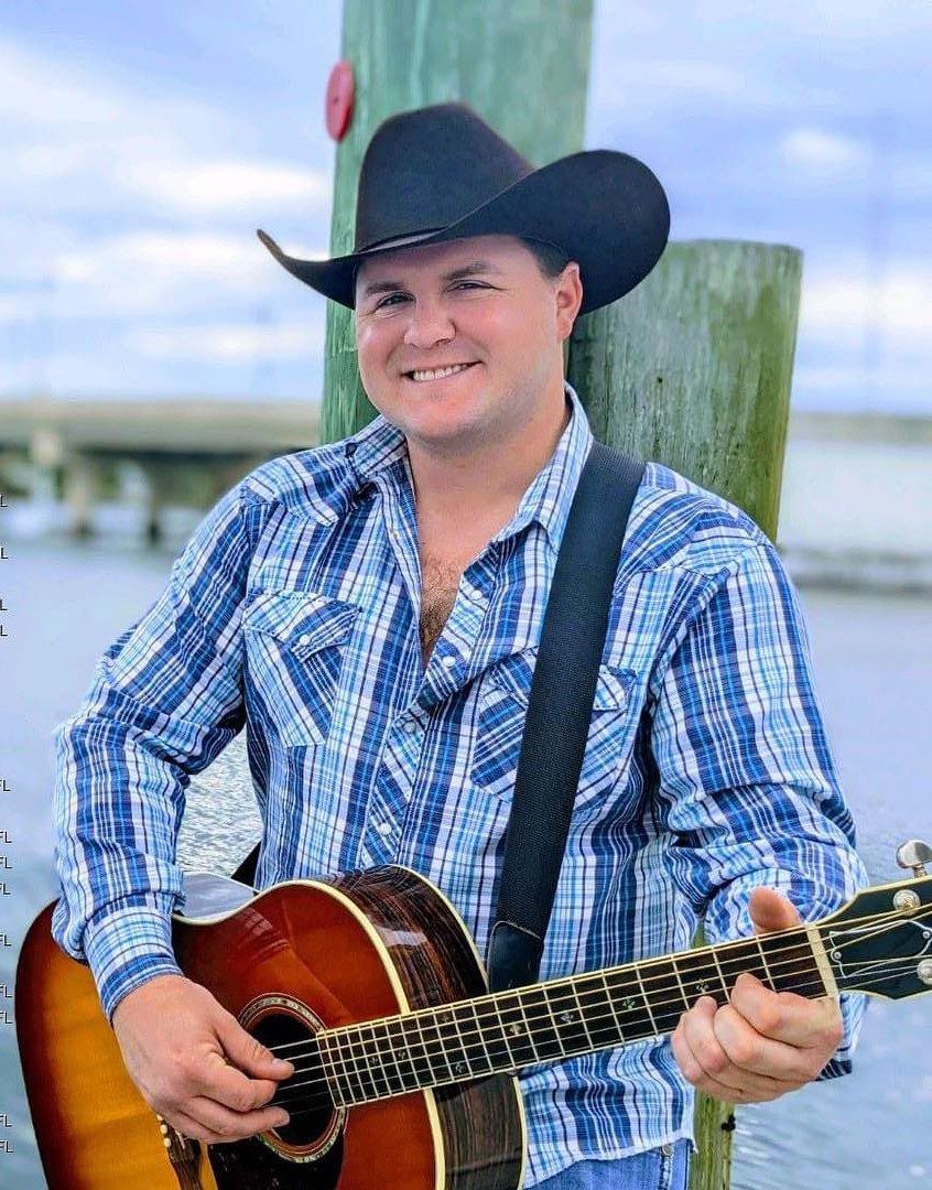 A man in cowboy hat playing guitar near water.