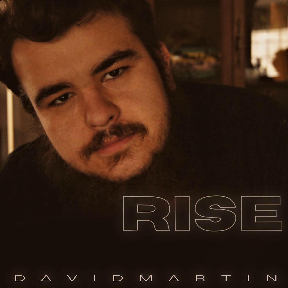 A man with a mustache and beard standing in front of the word rise.