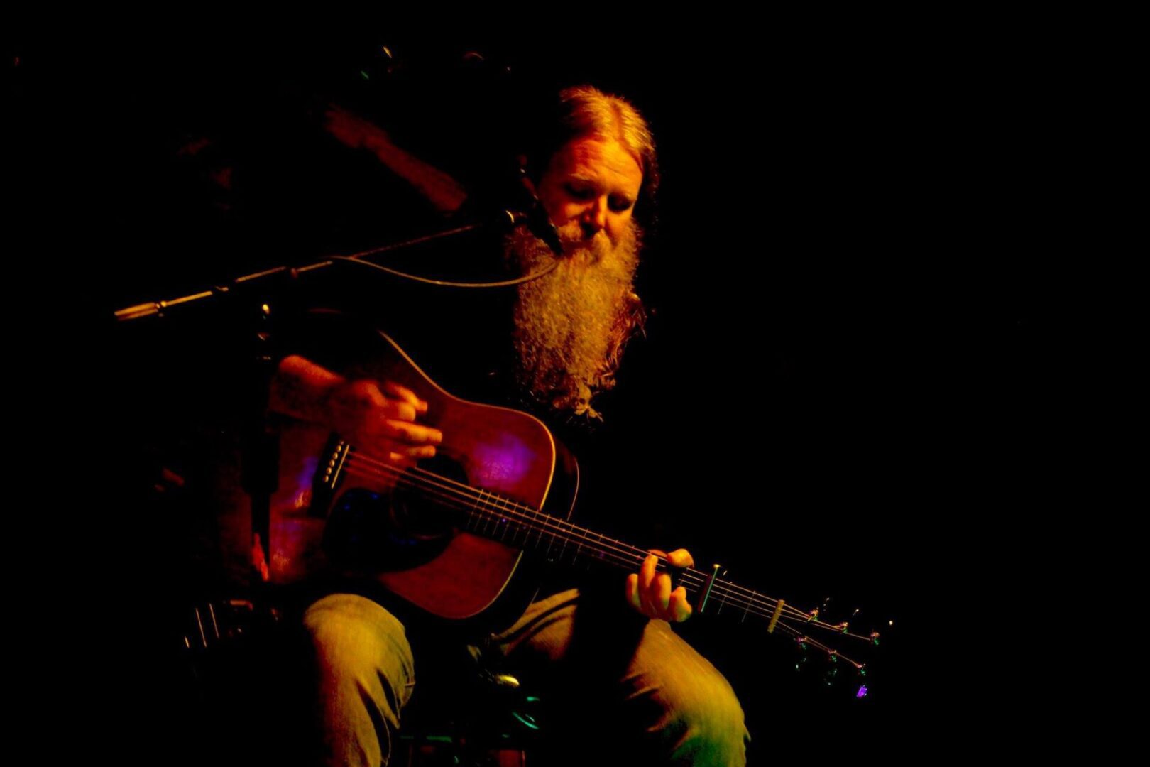 Albert Simpson: male with a mix of long brown and grey hair and a long beard; sitting down playing a guitar and is wearing a black shirt and jeans.