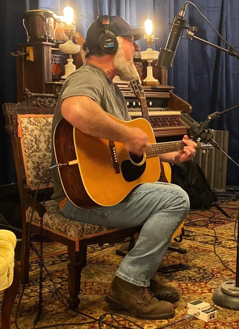 Michael Goldsmith sitting on a cushioned chair playing an acoustic guitar while wearing a grey T-shirt, light jeans, and brown boots.