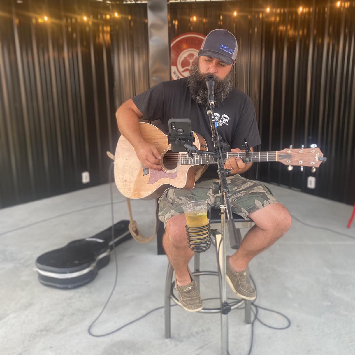 Mikey Wilson sitting on a stool waring a grey cap, black shirt, camo shorts, and brown shoes while singing and playing the acoustic guitar.