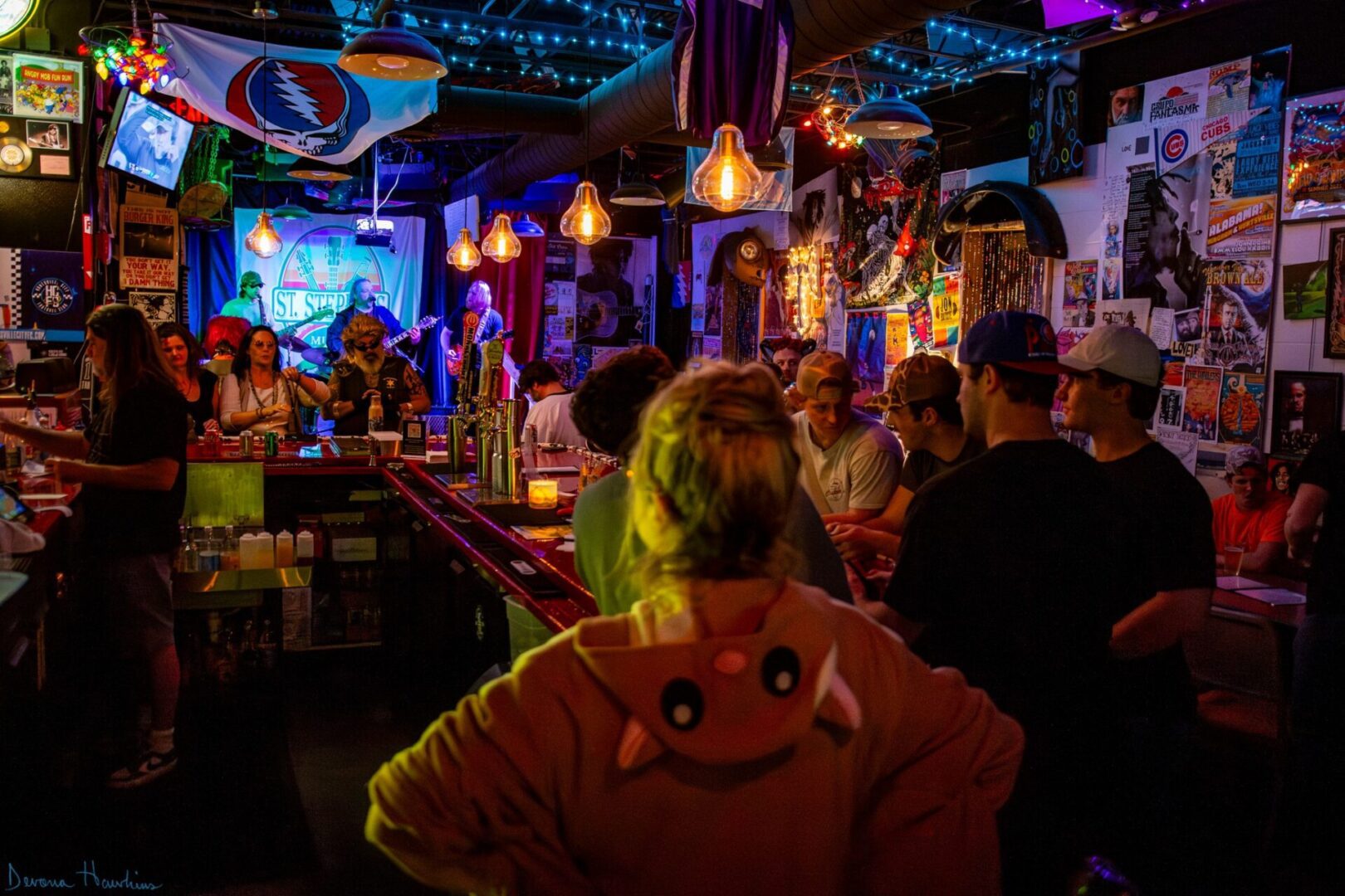 An inside look of St. Stephens Music Hall with a crowd off people sitting at the bar, posters plastered all on the walls, and a band playing on stage with a logo of the venue in the back.