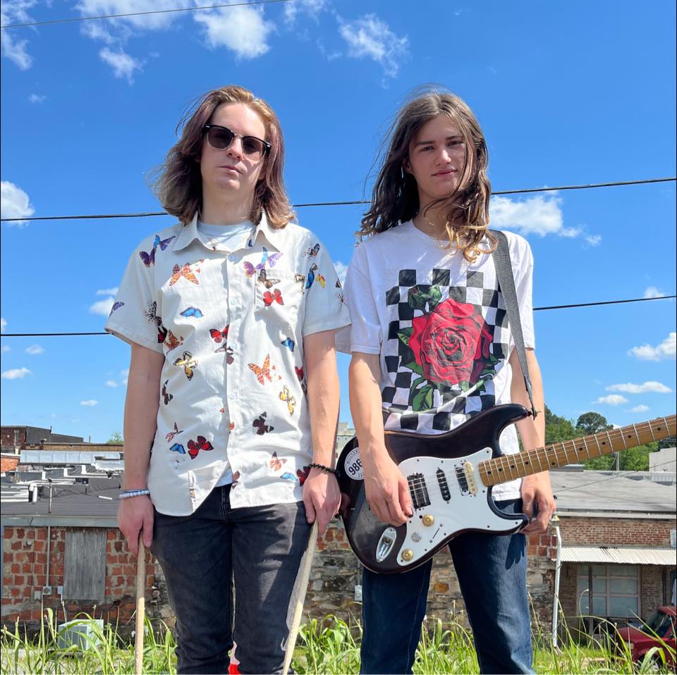 The two band members standing under a blue sky. Male member to the left had shoulder length brown hair, he is wearing sunglasses, a butterfly white button up shirt, and black pants. The member to the right has long brown hair, a white T-shirt with a rose on the front and a checkered pattern behind it, jeans, and he has an electric guitar slung over his shoulder.